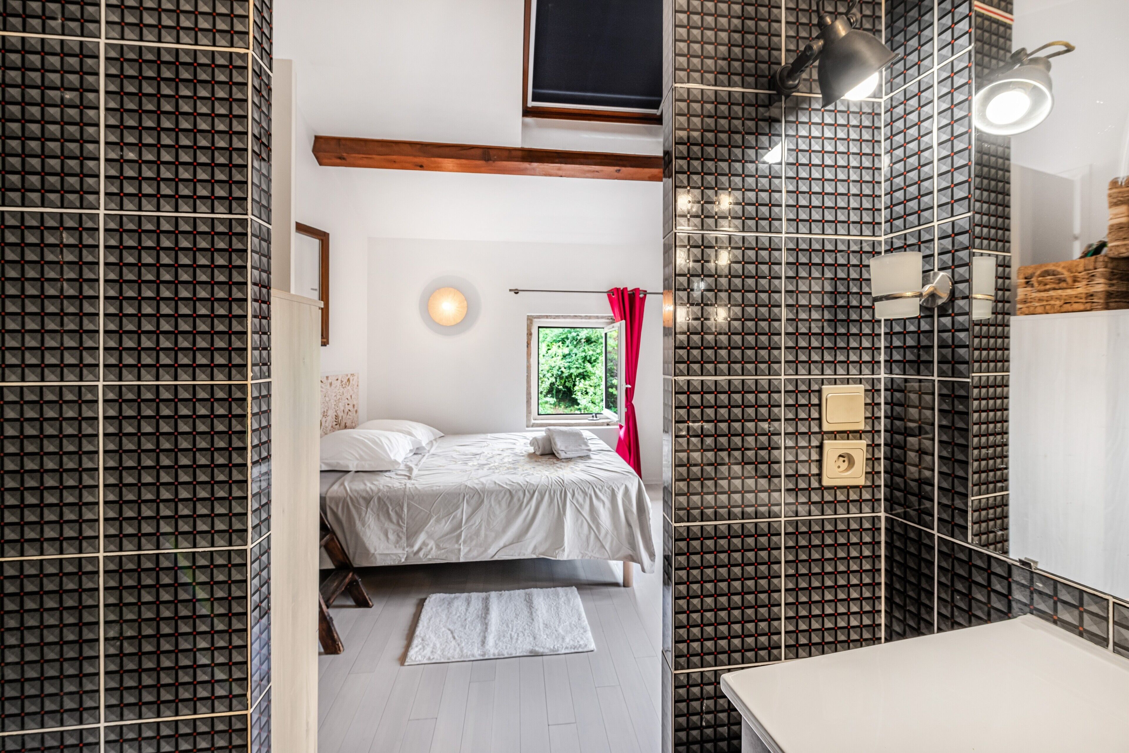 Un dormitorio moderno con cama, mesita de noche, ventana con vistas a zonas verdes, y baño con espejo y lavamanos.