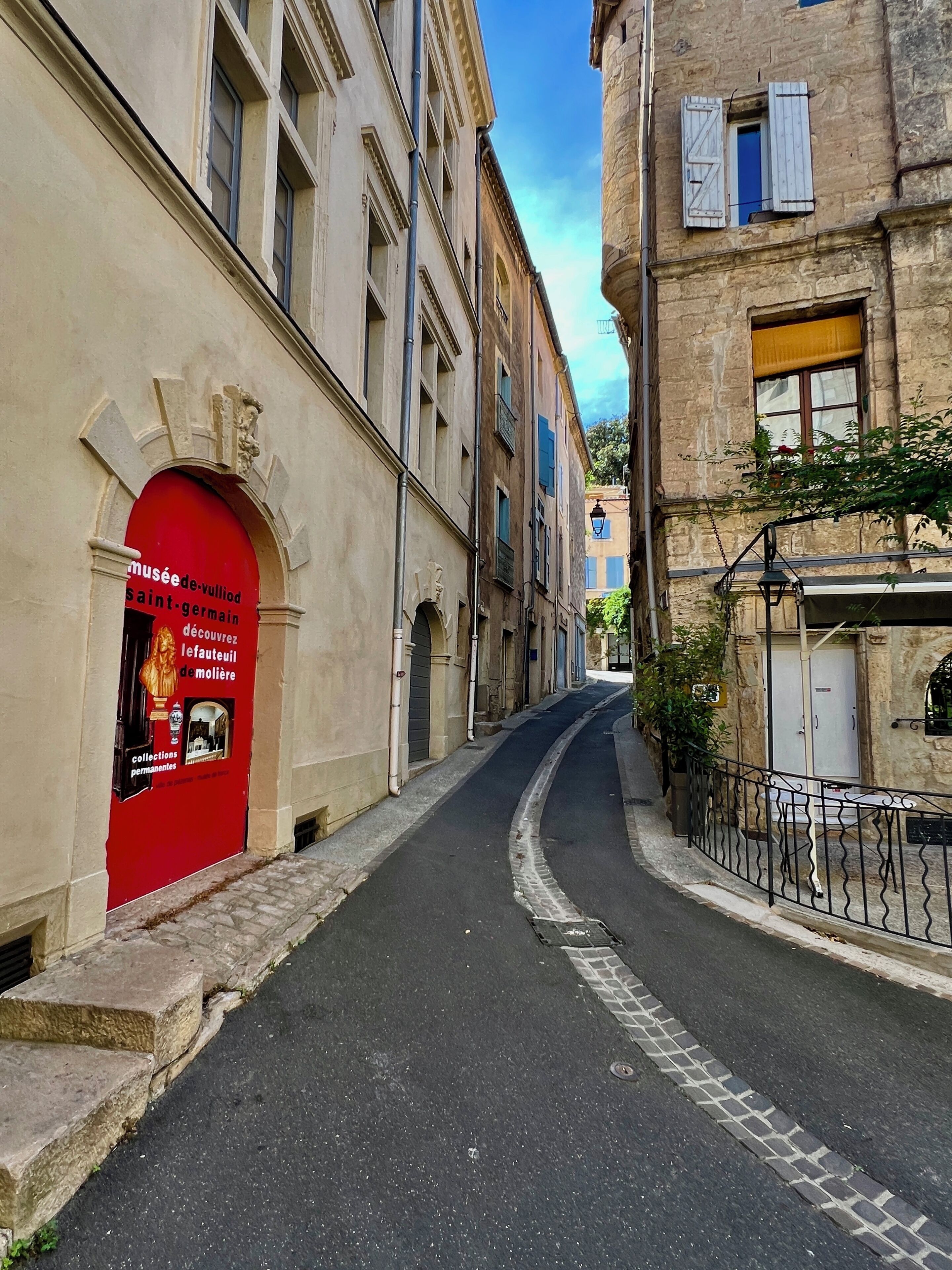 A narrow street with buildings on either side, a red door on the left, and a wrought iron fence on the right.