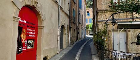 A narrow street with buildings on either side, a red door on the left, and a wrought iron fence on the right.