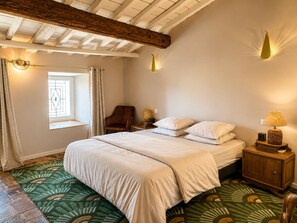 Ein Schlafzimmer mit einem Bett, einem Sessel, einem Nachttisch, einer Lampe, einem Fenster mit Vorhängen und einem gemusterten Teppich.