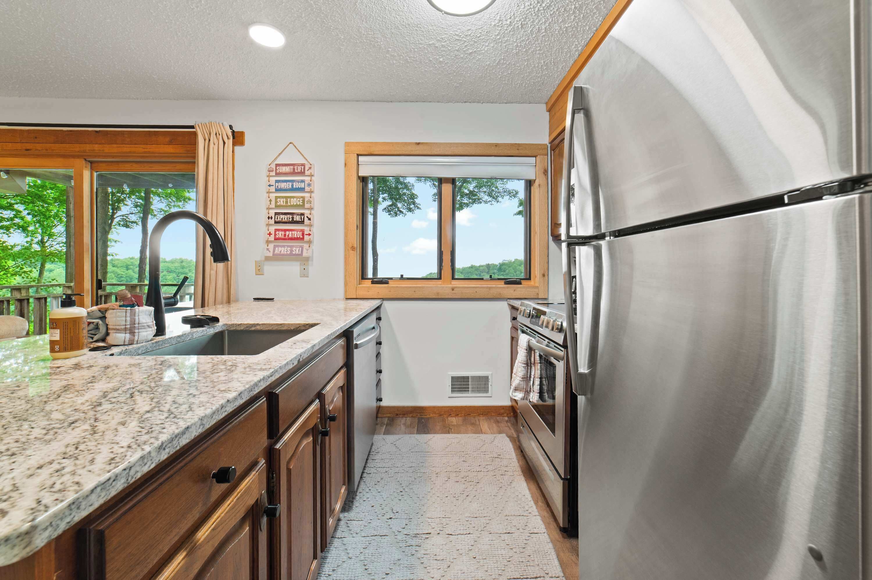 A modern kitchen with a stainless steel refrigerator, granite countertops, wooden cabinets, and a window with a view of trees.