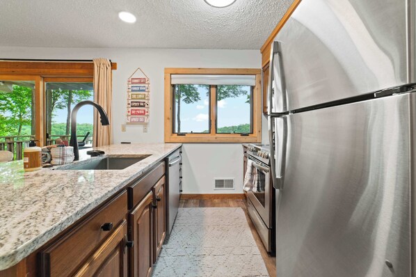 Cocina moderna con refrigerador de acero inoxidable, encimeras de granito, gabinetes de madera y una ventana con vista a árboles.