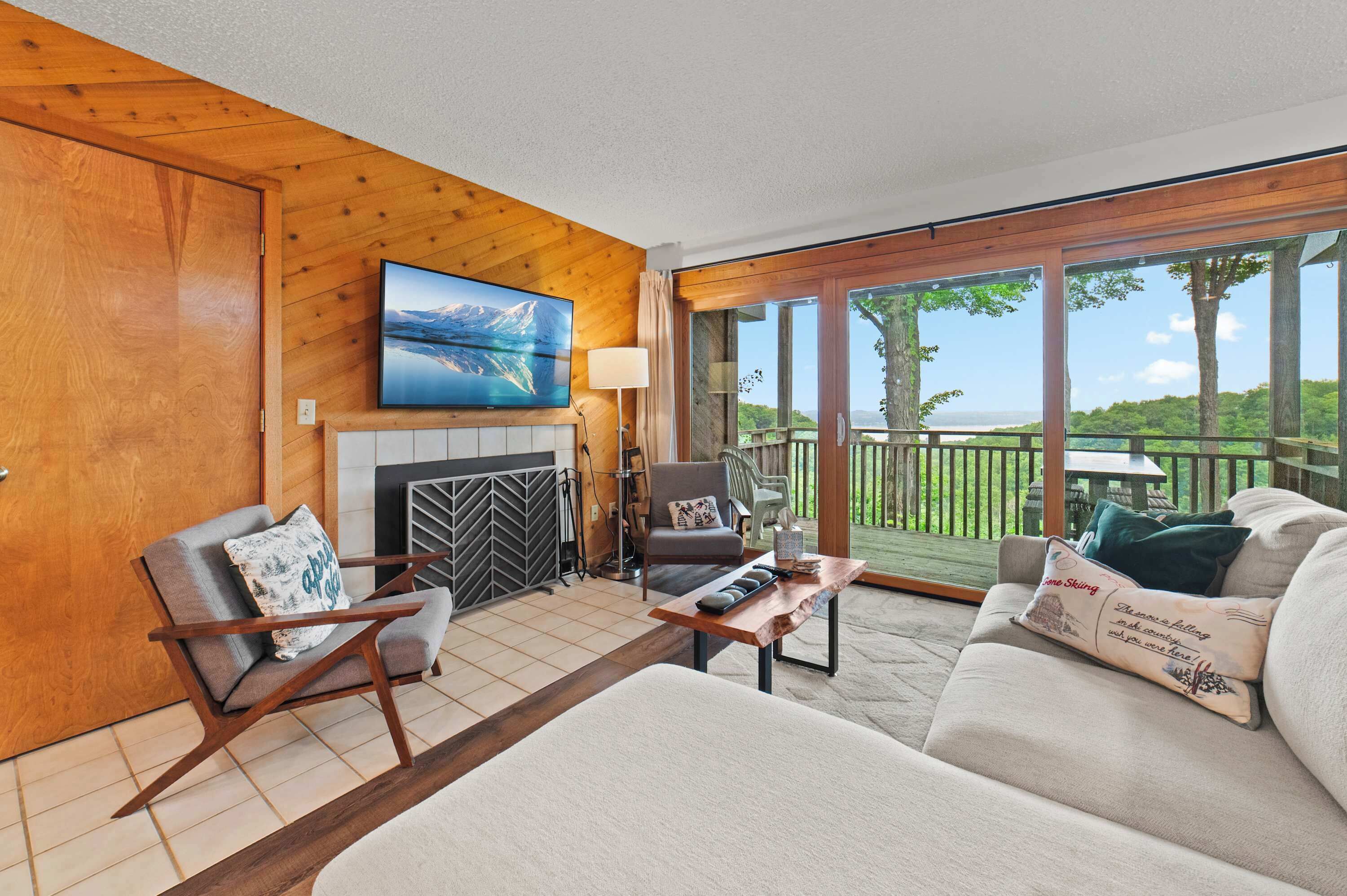 A living room with a fireplace, a sofa, a chair, and a table. There is a large window with a view of trees and a deck.