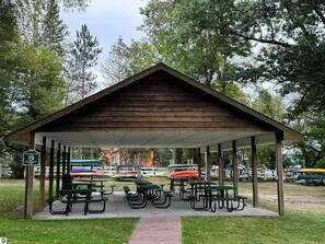 A covered picnic area with benches and picnic tables, surrounded by trees. - Lake Bellaire View Condo | Pool • Beach Club (Bellaire)