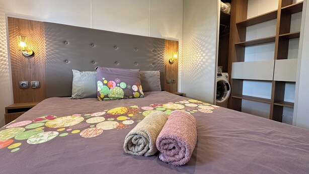 A neatly made bed with a floral bedspread, two rolled towels, and a headboard with wall-mounted lights.