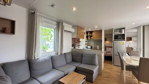 A modern living room with a grey sectional sofa, a wooden coffee table, and a kitchen area with a microwave, refrigerator, and wooden cabinets.