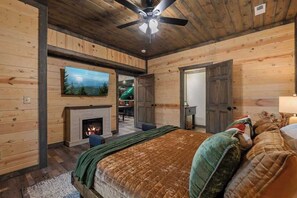 A log cabin interior with a fireplace, a bed with multiple pillows, a ceiling fan, and a television mounted on the wall. - 8-Bedroom Cabin with Themed Indoor Pool, Theater & Arcade Near Pigeon Forge (Sevierville)