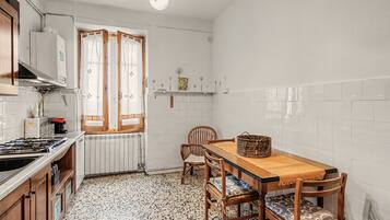 Eine kleine Küche mit Gasherd, Holzschränken, Fliesenboden, einem Esstisch mit Stühlen und einem Fenster mit Vorhängen.