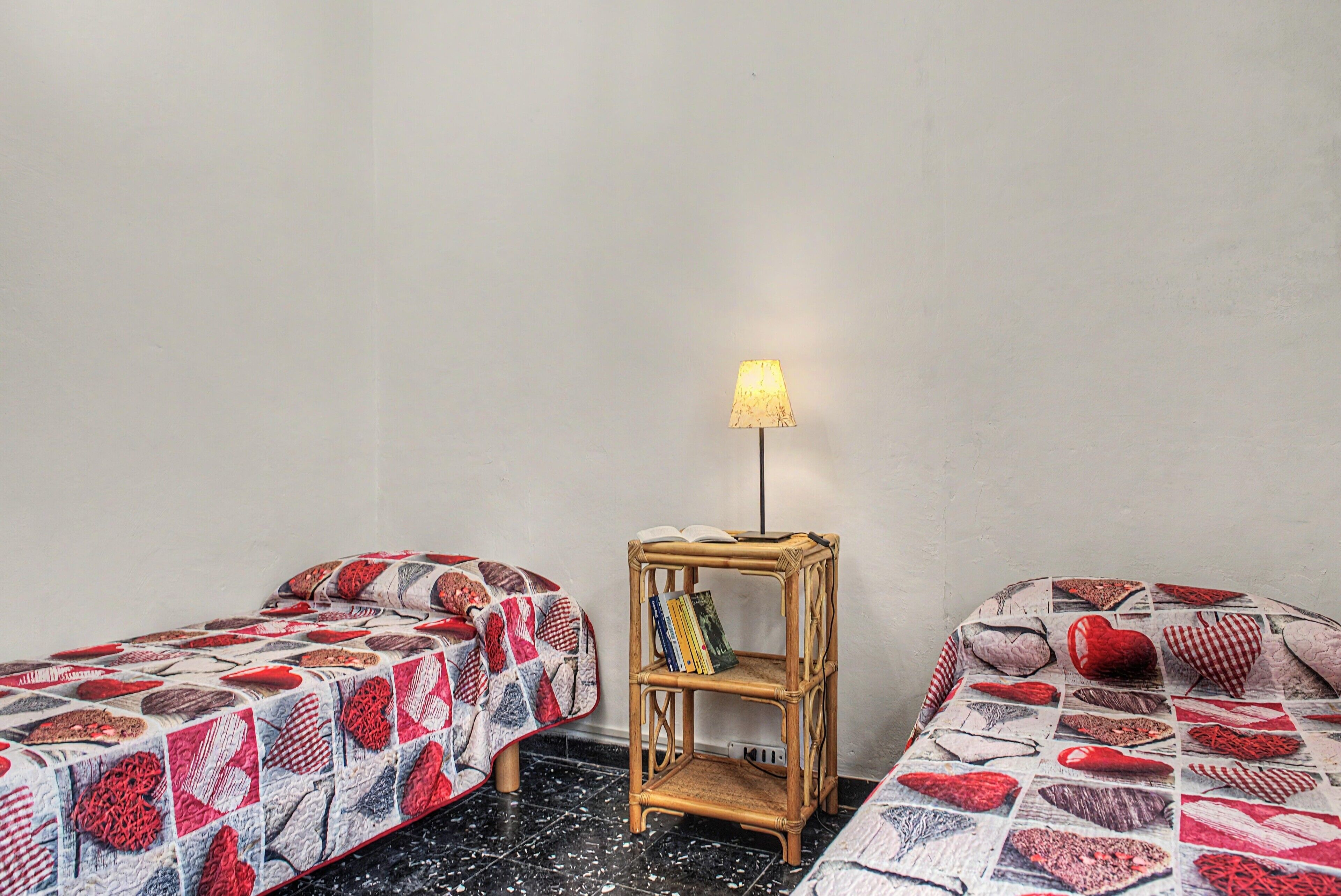 Ein kleines Zimmer mit zwei Betten, einem Nachttisch mit Lampe und einem an der Wand befestigten Regal mit Büchern.