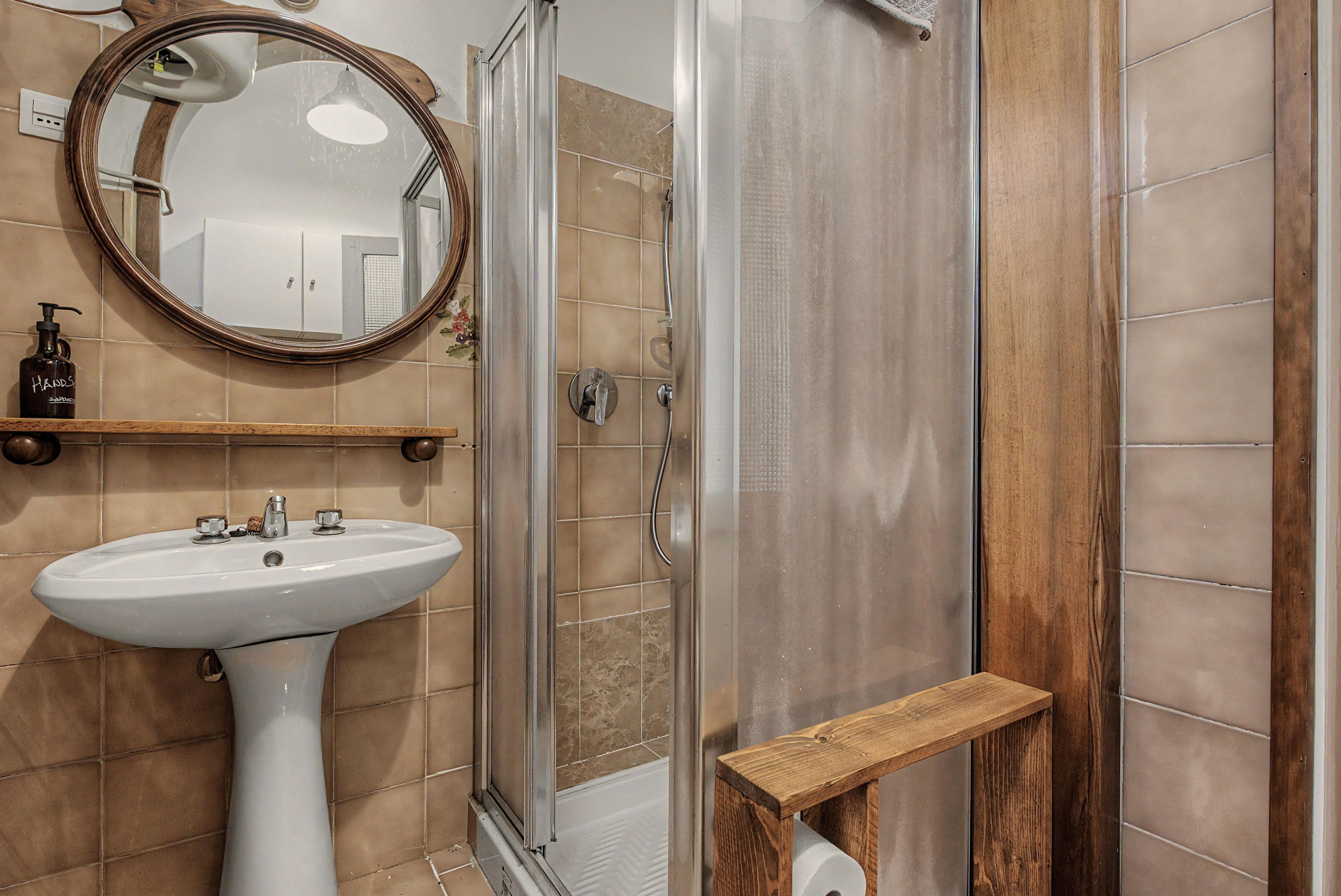 Une salle de bain avec un lavabo sur colonne, un espace douche, une étagère en bois et un miroir.