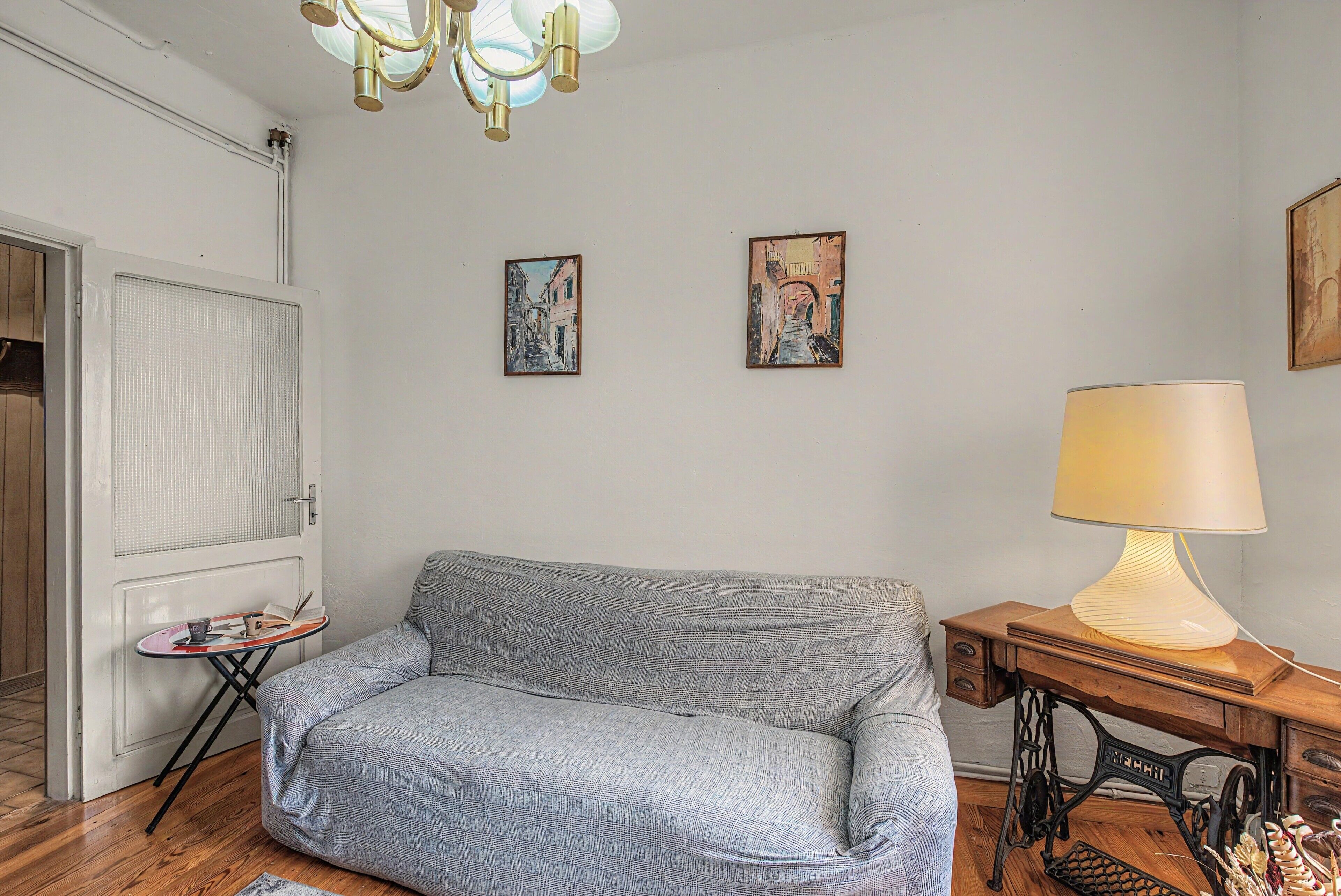 A room with a patterned sofa, a small table with a lamp, and two paintings on the wall.