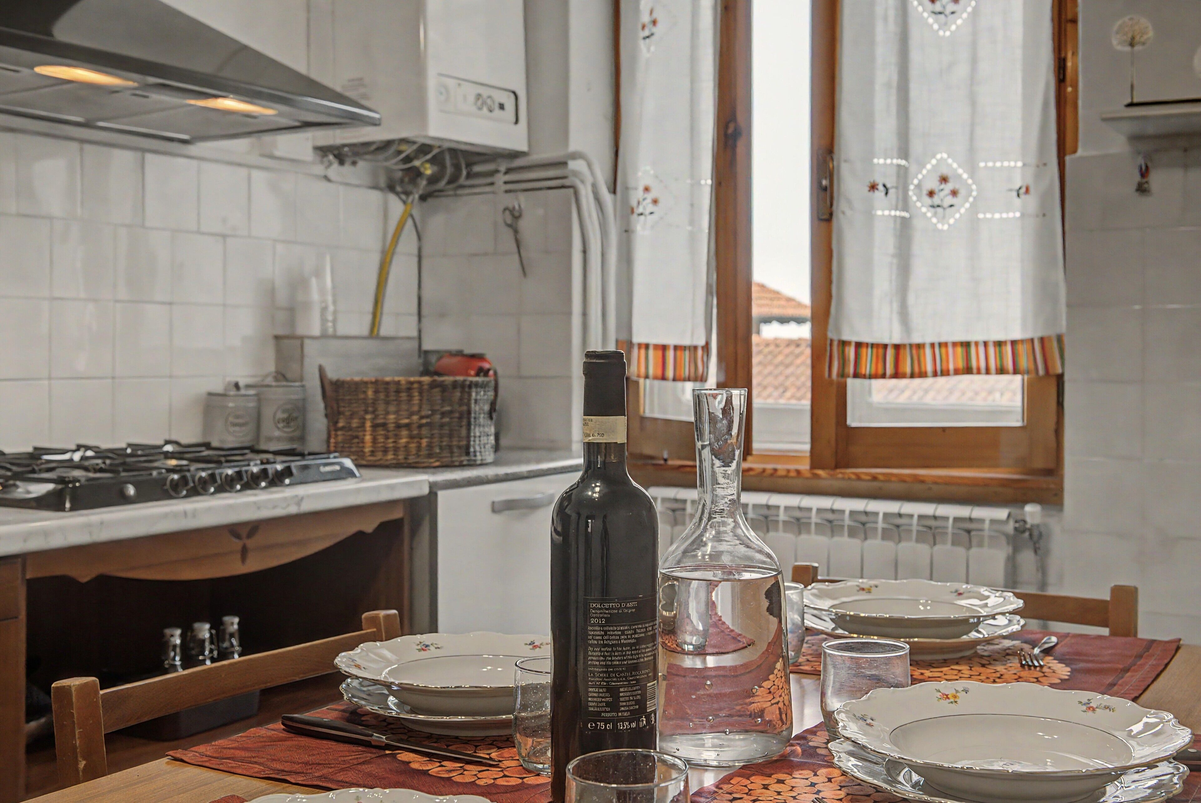 A kitchen with white tiles, a gas stove, a bottle, a glass carafe, and a dining table set with plates and cutlery.