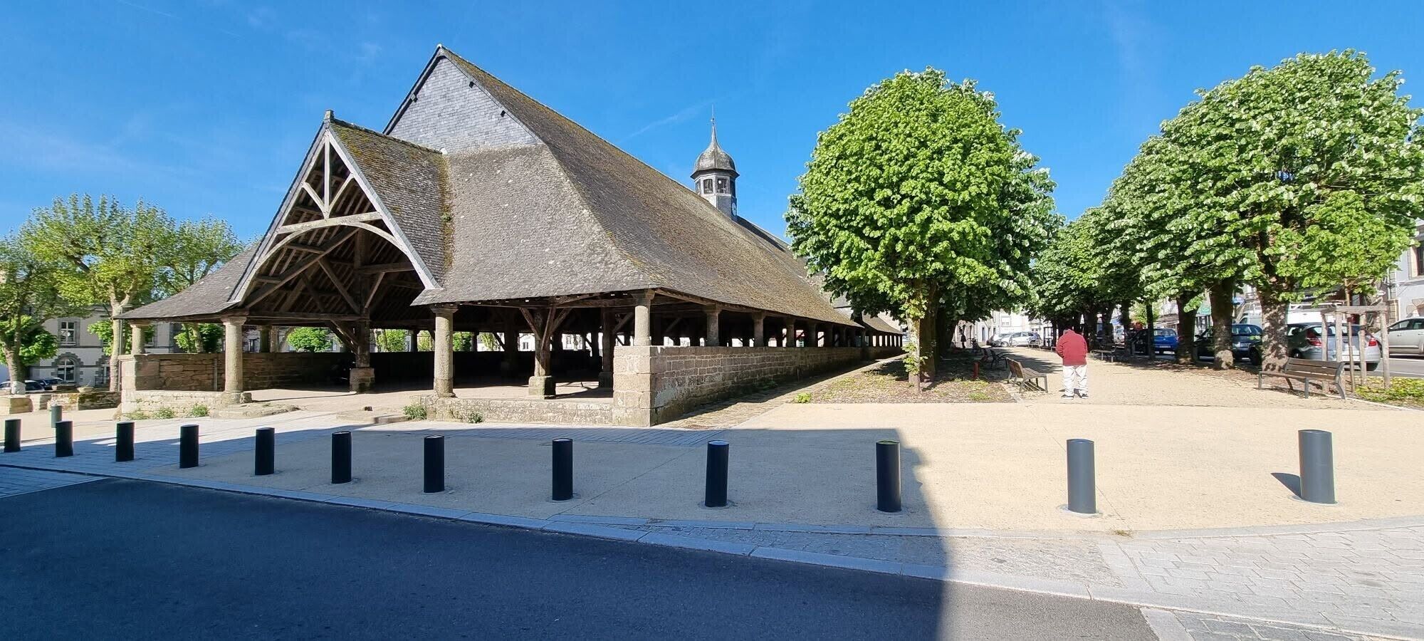 A historical building with a triangular roof and a clock tower, surrounded by trees and a paved area with bollards.