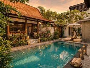 Un espace piscine doté d’une terrasse en bois, de chaises longues et d’un coin salon couvert avec des parasols.
