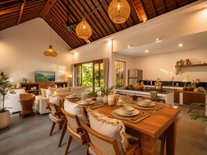 Een ruime eethoek met een houten tafel voor vier personen, omringd door rieten stoelen. Achtergrond toont een keuken met moderne apparatuur en een groot raam.