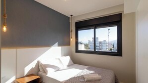 A modern bedroom with a bed, a nightstand, a window with a view of buildings, and a hanging light fixture. - Apt in Condo w/ Private BBQ in Center Ilg0307 (Bombinhas)