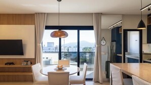A modern hotel room with a dining area, a flat-screen TV, a large window with a view of buildings, and a dining table with chairs. - Apt in Condo w/ Private BBQ in Center Ilg0307 (Bombinhas)