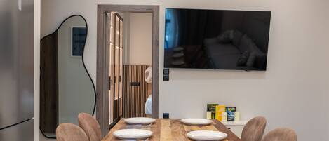 A dining area with a wooden table set for six, a television mounted on the wall, and a door leading to another room.
