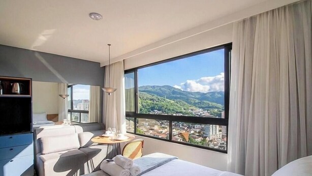 A hotel room with a large window, a bed, a seating area, and a desk. - Studio in Condo w/ Pool in Blumenau Pin1906 (Blumenau)