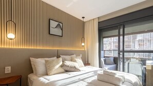 Ein modernes Hotelzimmer mit einem großen Bett, Blick auf die Stadt von einem Balkon und einem an der Wand befestigten Kunstwerk.
