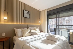 Ein modernes Hotelzimmer mit einem großen Bett, Blick auf die Stadt von einem Balkon und einem an der Wand befestigten Kunstwerk.