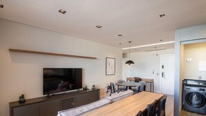Ein modernes Wohnzimmer mit Flachbildfernseher, einem Essbereich mit Holztisch und einer Waschmaschine in der Ecke.