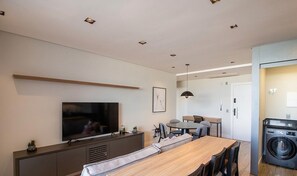 A modern living room with a flat-screen TV, a dining area with a wooden table, and a washing machine in the corner. - Condo Apt w/ Pool in Estados, Balneario Tso0314 (Balneário Camboriú)