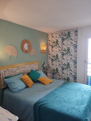 Ein Bett mit gemusterten Textilien, ein Vorhang mit dekorativem Design, ein Wandventilator, eine Wandleuchte und floraler Wandtapete.