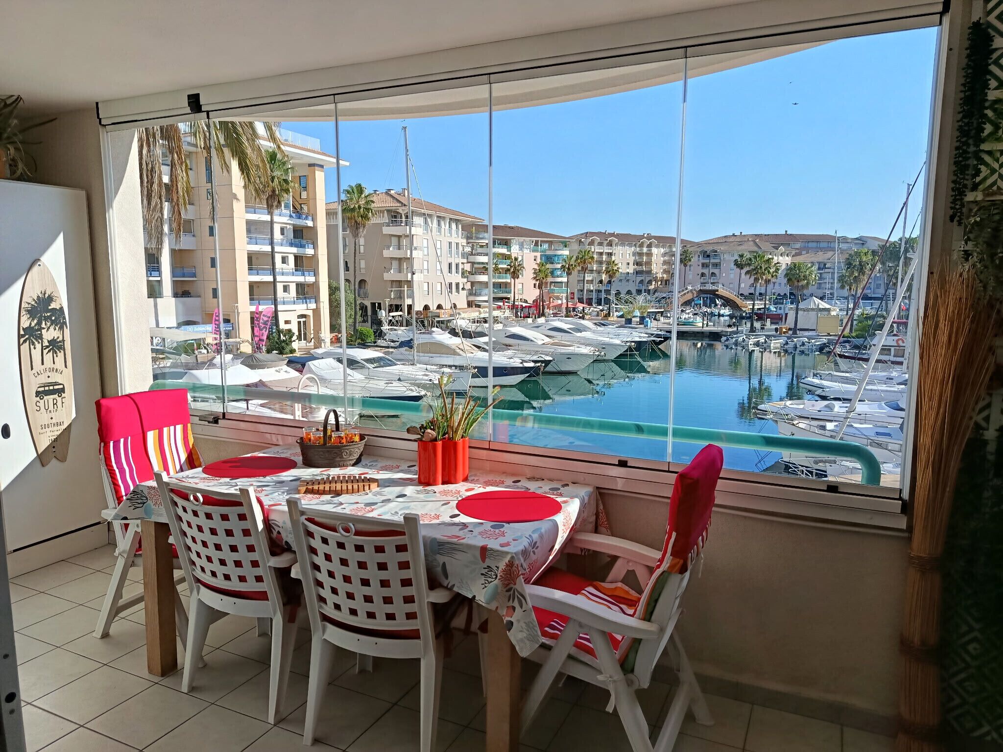 A dining area with a table and chairs, a view of a marina with boats, and a building with balconies in the background.