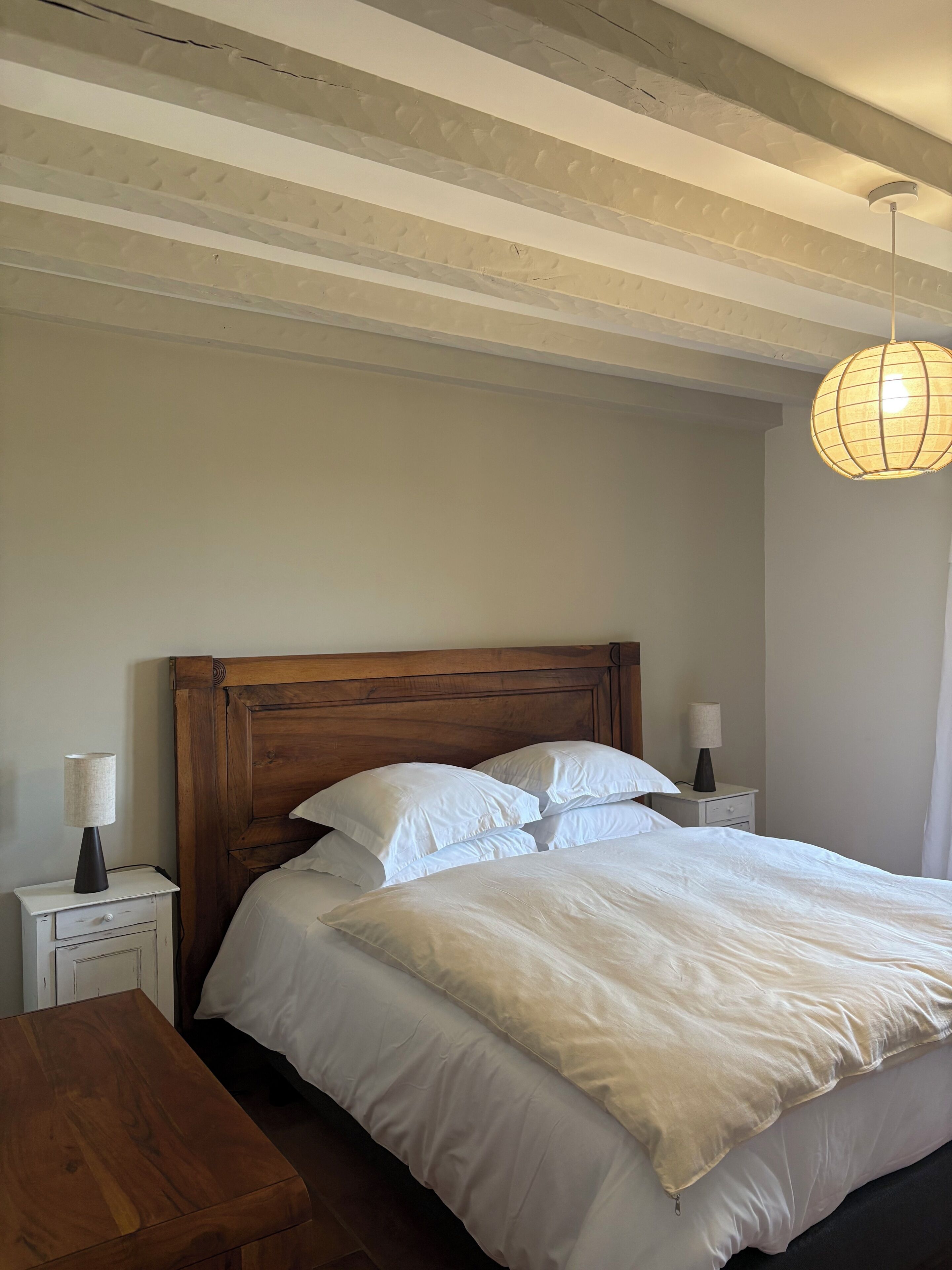 Une chambre à coucher avec une tête de lit en bois, un lit recouvert de draps blancs, deux tables de chevet avec des lampes et un luminaire suspendu.