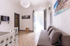 Ein Wohnzimmer mit einer Couch, einem an der Wand montierten Fernseher, einer Tür und einem Fenster mit durchsichtigen Vorhängen.
