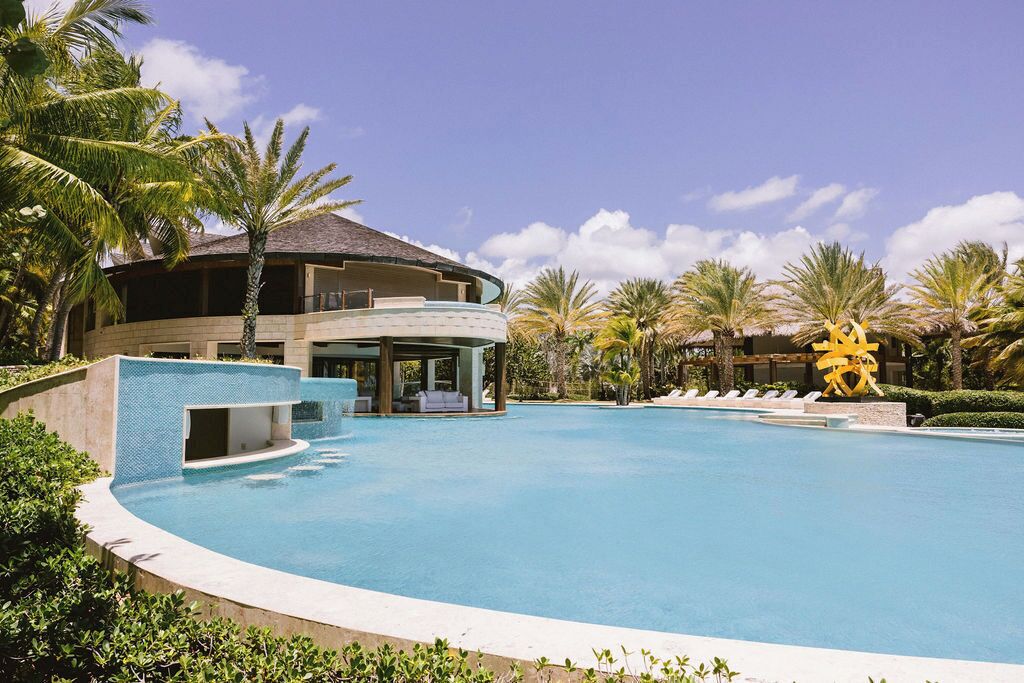 Een resort met een groot zwembad, palmbomen en een opvallend geel sculptuur.