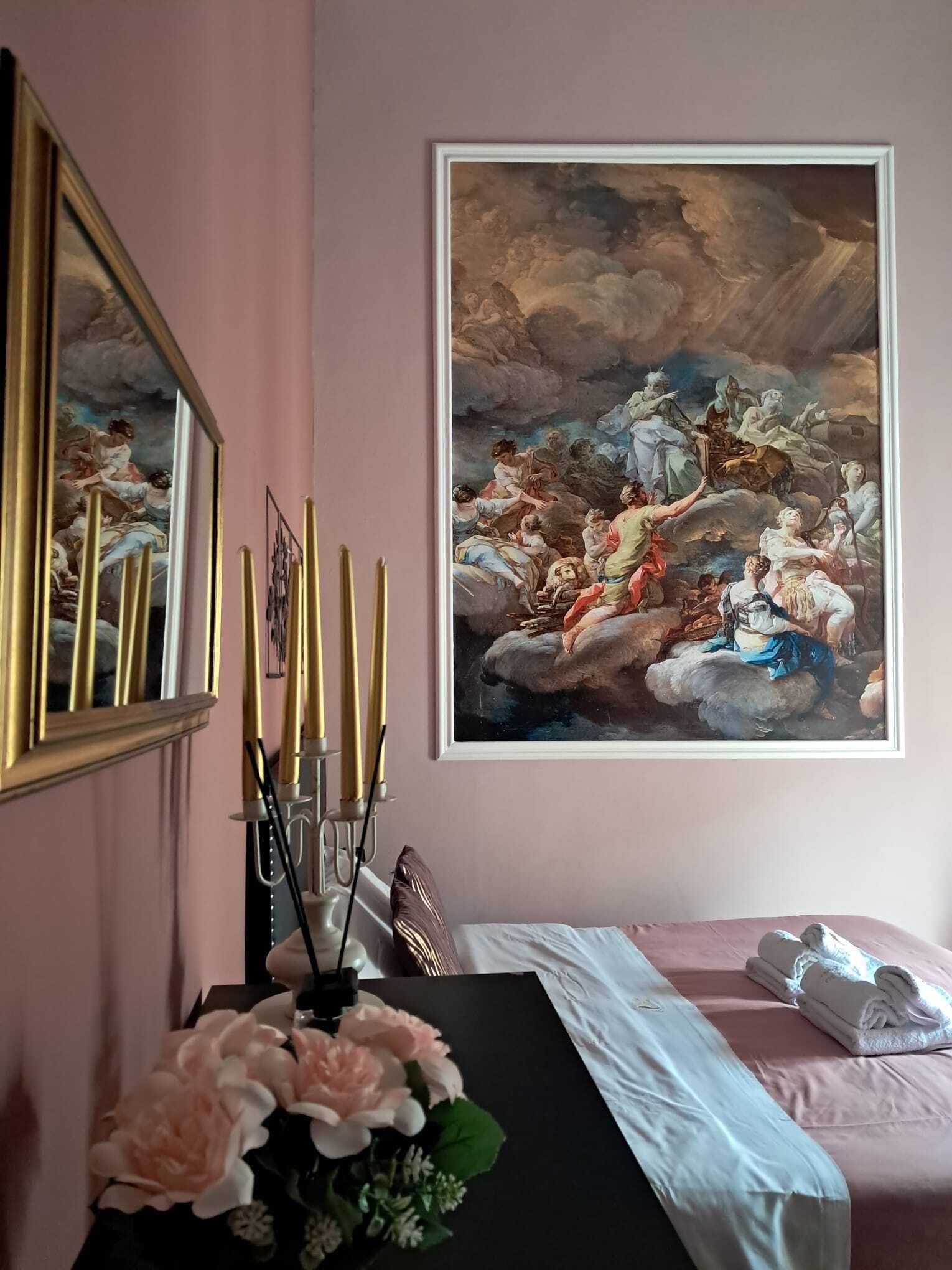 Ein Zimmer mit einem großen Gemälde im klassischen Stil, einem Spiegel, einem Nachttisch mit Kerzen und einem Bett mit rosa Bettdecke.