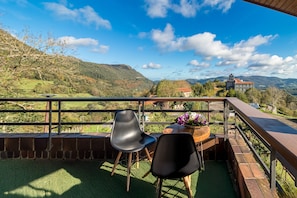 A balcony with two chairs and a table with flowers, overlooking a mountainous landscape and a church.