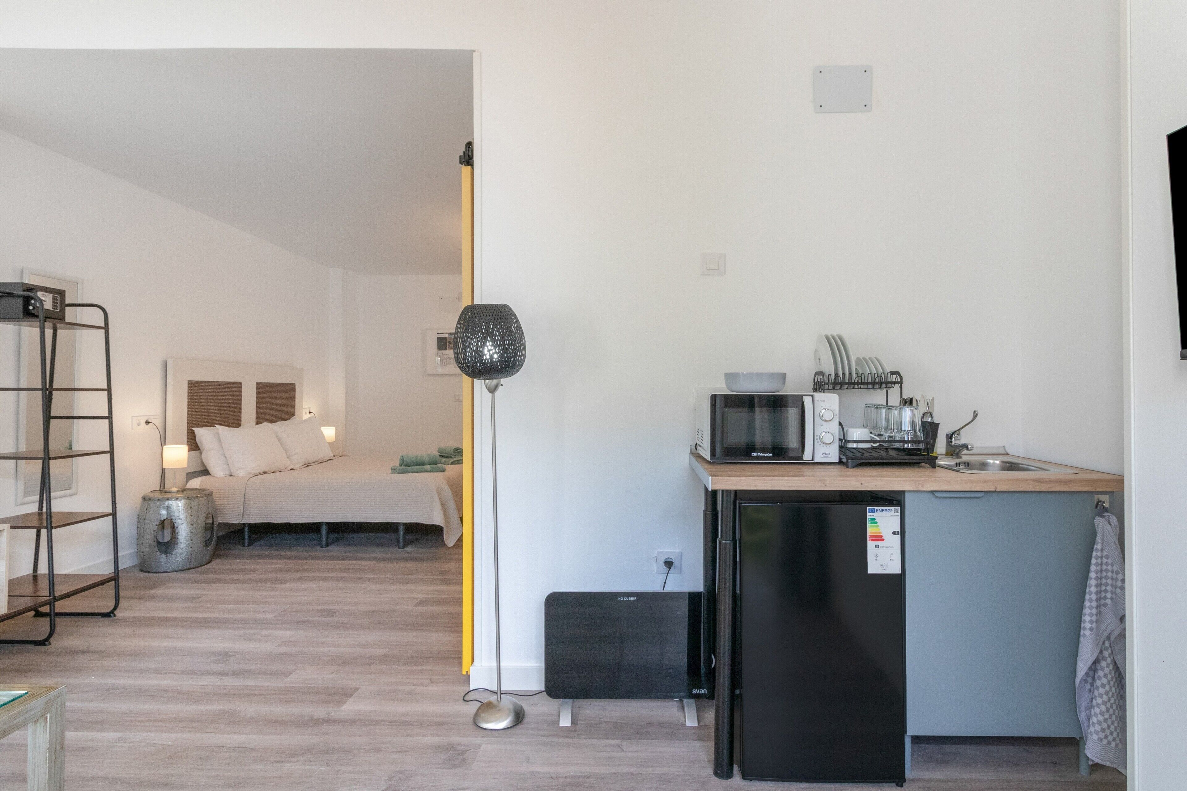 A compact hotel room with a kitchenette, a bed with pillows, a standing lamp, and a small side table.