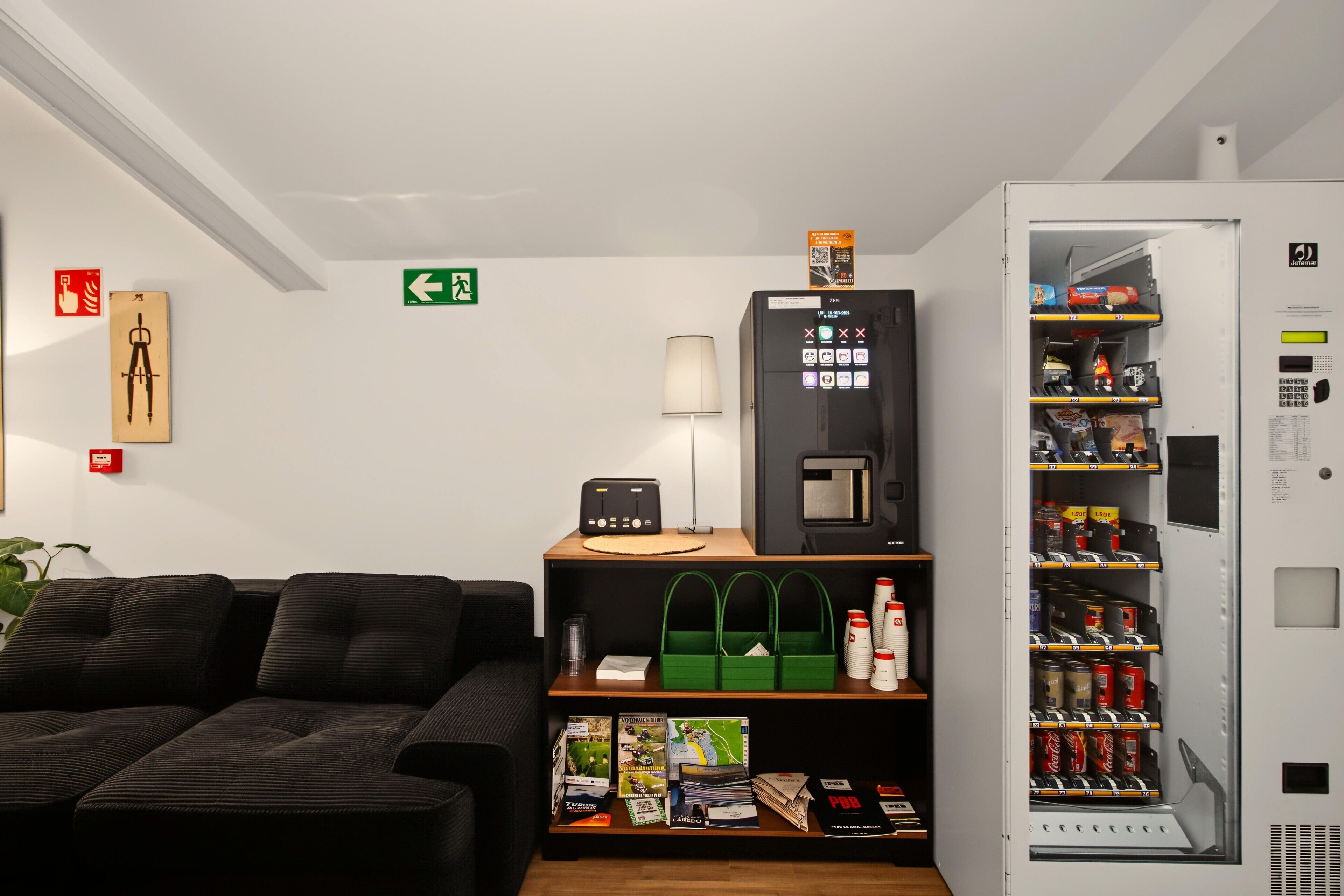 Habitación de hotel con sofá, una mesita con lámpara, una máquina expendedora y un estante con varios objetos.