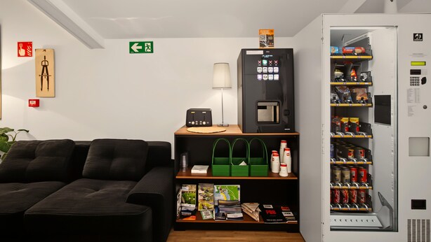 Habitación de hotel con un sofá negro, una mesita con una lámpara, una máquina expendedora y un estante con revistas y una bolsa verde.