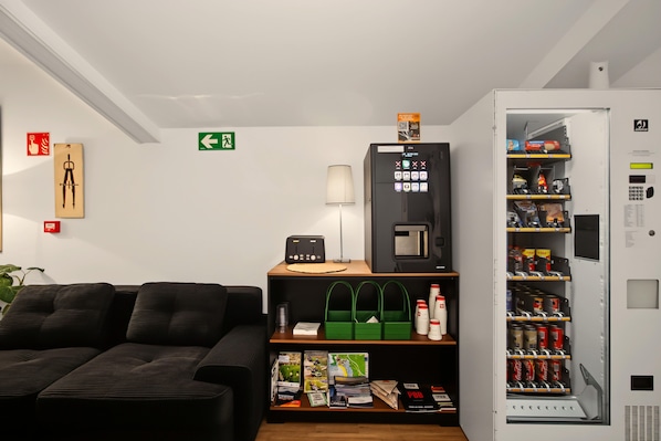 A hotel room with a sofa, a small table with a lamp, a vending machine, and a shelf with magazines and a green bag.