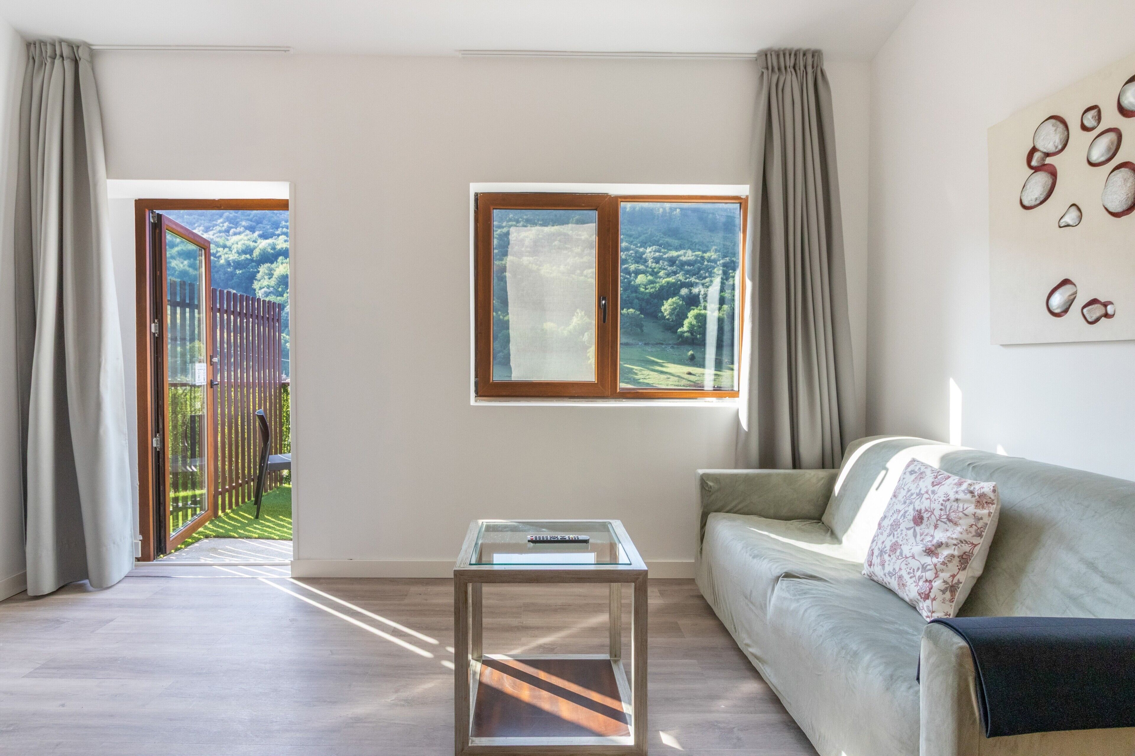 Una sala de estar con un sofá, una mesa de centro de vidrio y dos ventanas con vistas a un jardín y árboles.