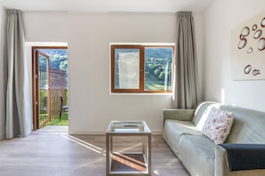 A living room with a sofa, a glass coffee table, and two windows offering views of a garden and trees.