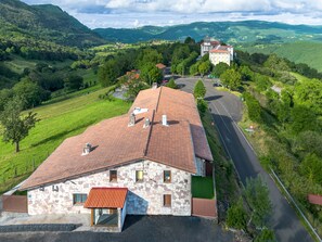 A stone building with a red-tiled roof, a parking area, and a view of a road leading to a hilltop church.