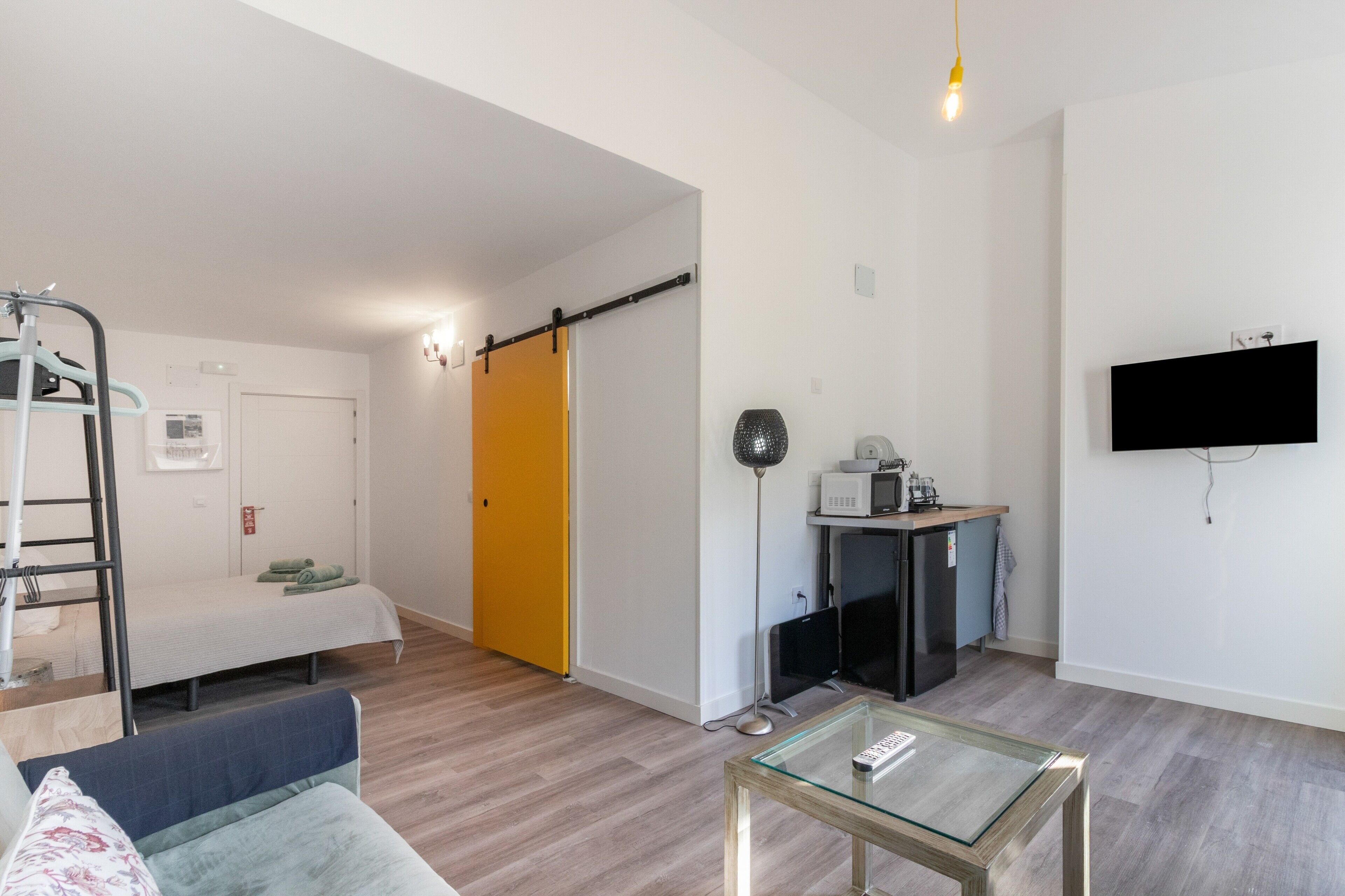 Un salon moderne comprenant un canapé, une table basse en verre, un téléviseur fixé au mur, un coin cuisine avec un four à micro-ondes et une porte coulissante donnant sur une autre pièce.