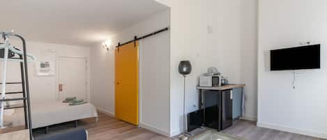 Una sala moderna con un sofá, una mesa de centro de vidrio, un televisor montado en la pared, una zona de cocina con microondas y una puerta corredera que da a otra habitación.