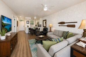 A living room with a sofa, armchair, coffee table, and a TV. There is a dining area with chairs and a table. The kitchen is visible in the background. - Oceanfront Maui Condo, Whale Watching, Beach Access & Endless Views (Wailuku)