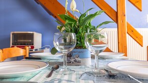 A dining table with two wine glasses, a plate, and a spoon. There is a potted plant with green leaves and a white flower. The table is set against a blue wall with wooden beams. - Duplex No. 2345 – 40m² – Seaside Residence (Port-des-Barques)