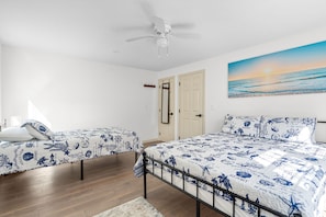A bedroom with two beds, a ceiling fan, a door, and a painting of a beach scene. - Mashpee Gem with a Private Yard, Volleyball, BBQ and a Fire pit! (Mashpee)