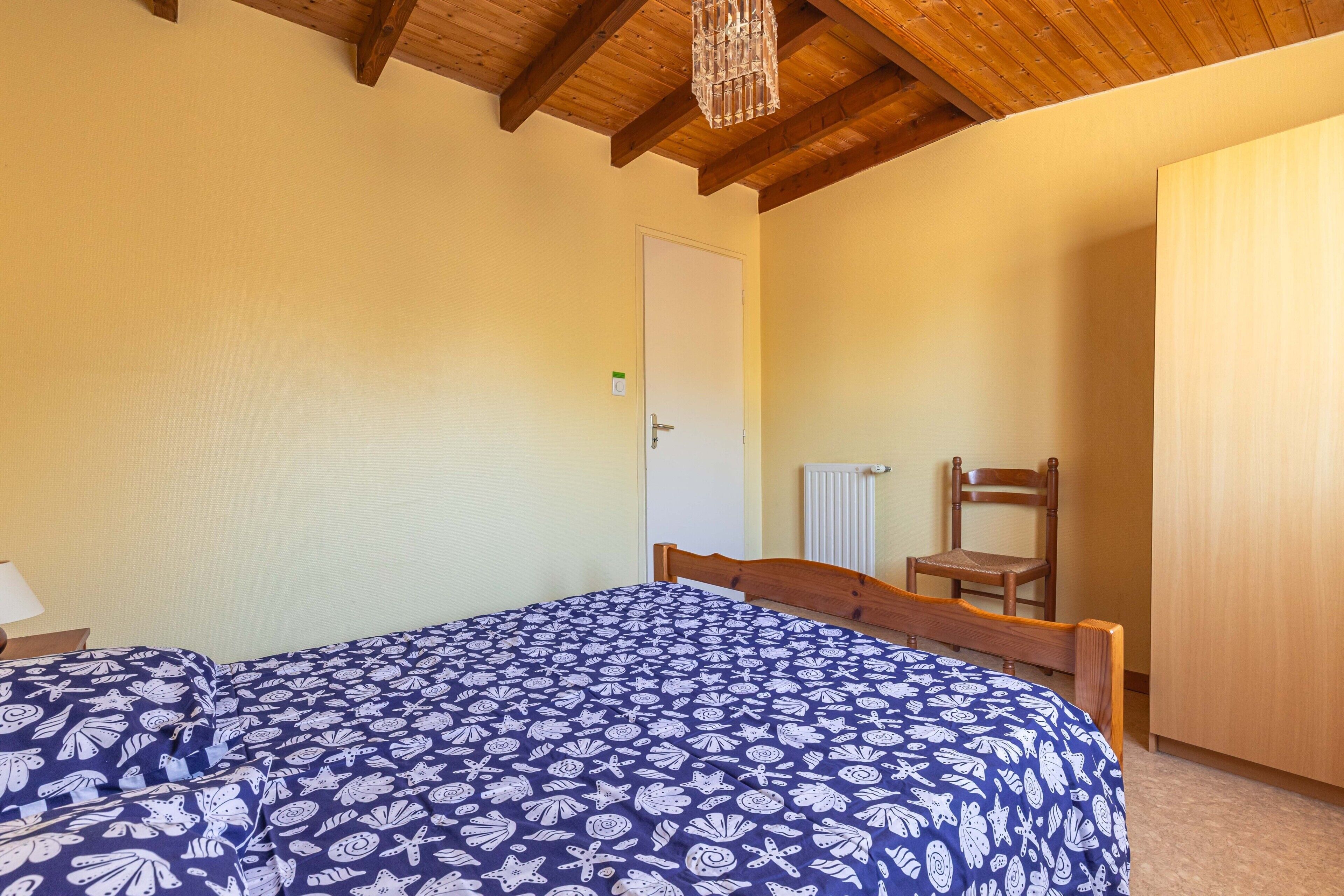 Un dormitorio con techo de madera, una cama con colcha estampada, una silla y un radiador.
