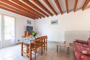 Ein Essbereich mit Tisch für zwei Personen, Holzstühle, ein kleiner Tisch mit einer Karaffe und eine Couch mit gemusterten Kissen.
