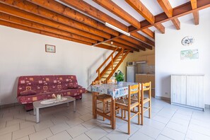A cozy living room with a wooden ceiling, a sofa, a dining table with chairs, a kitchen area with a refrigerator, and a staircase. - Duplex No. 1 – 70m² – Seaside Residence (Port-des-Barques)