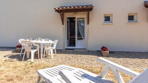 Ein Sitzbereich im Freien mit Tisch und Stühlen, eine überdachte Terrasse und ein Gebäudeeingang.
